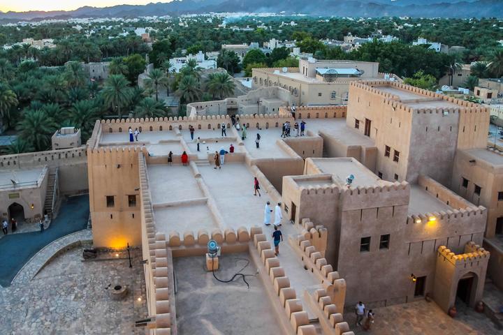Rooftops and walls of Nizwa Fort at dusk, with palm groves and mountains behind